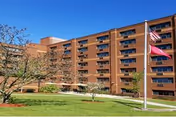 Brick multi-story senior living building with balconies, a manicured lawn, walkway, and two flagpoles under a clear blue sky.
