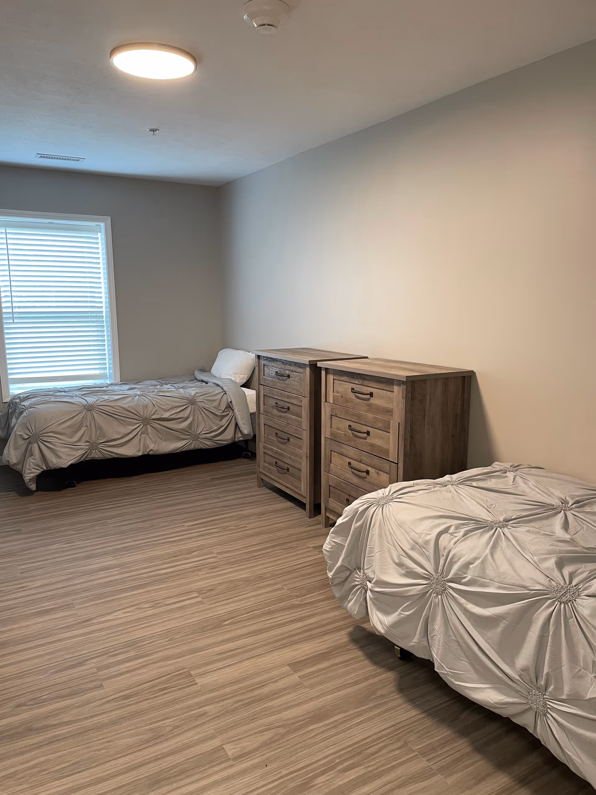 A simple bedroom with two twin beds, each covered with a light gray textured comforter. Between the beds are two wooden dressers with multiple drawers. A window with closed blinds is on the left wall, and the floor is covered with light wood laminate. The walls are painted a neutral beige color.