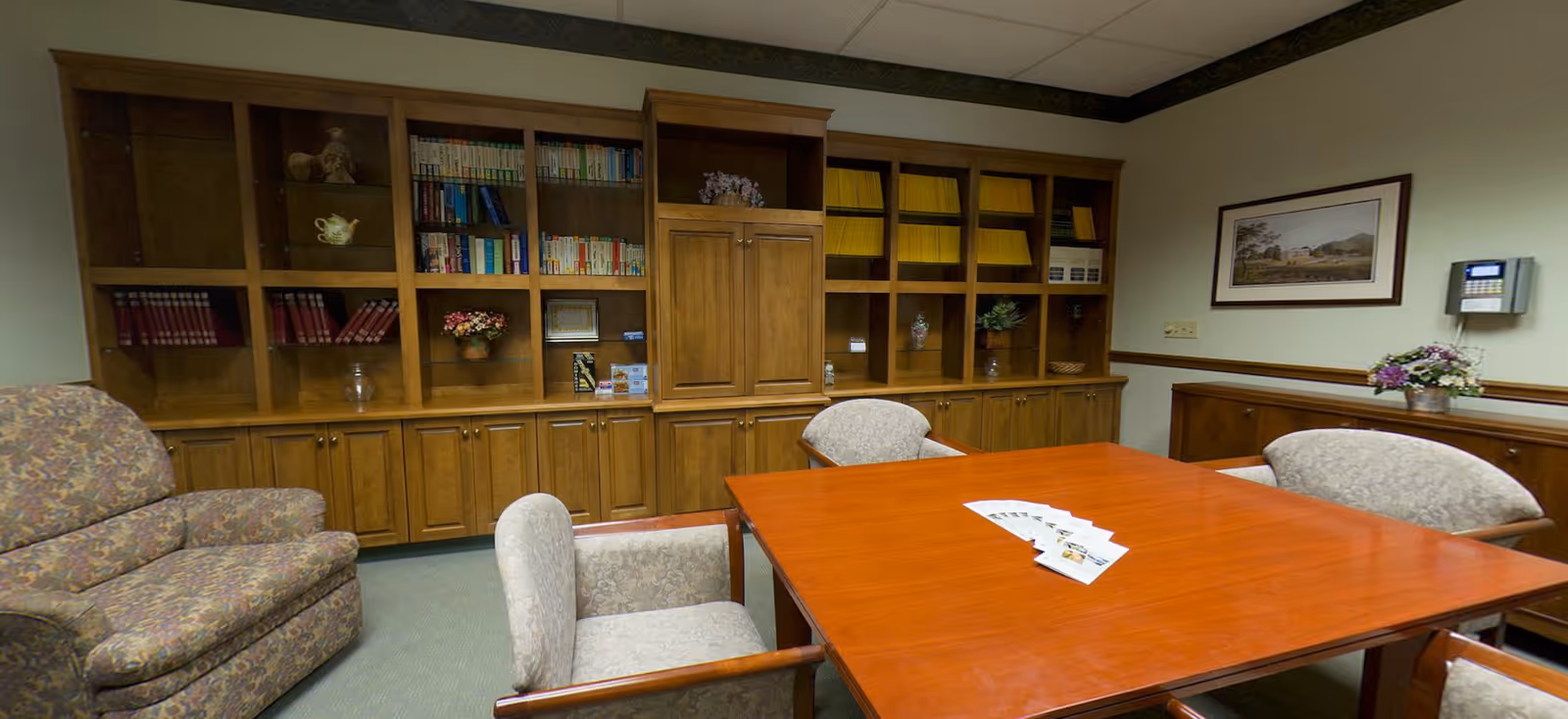 A meeting or conference room with a large wooden table surrounded by upholstered chairs. Behind the table is a wall with built-in wooden shelves containing books, magazines, and decorative items. There is a floral-patterned armchair to the left and a framed picture on the wall to the right, along with a small floral arrangement on a side cabinet.