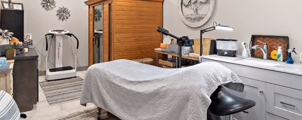 A wellness or therapy room with a massage table covered by a gray quilted blanket, a black stool, a wooden sauna cabin, a white vibration plate machine, and a countertop with a sink, lamp, and various wellness equipment and decor.