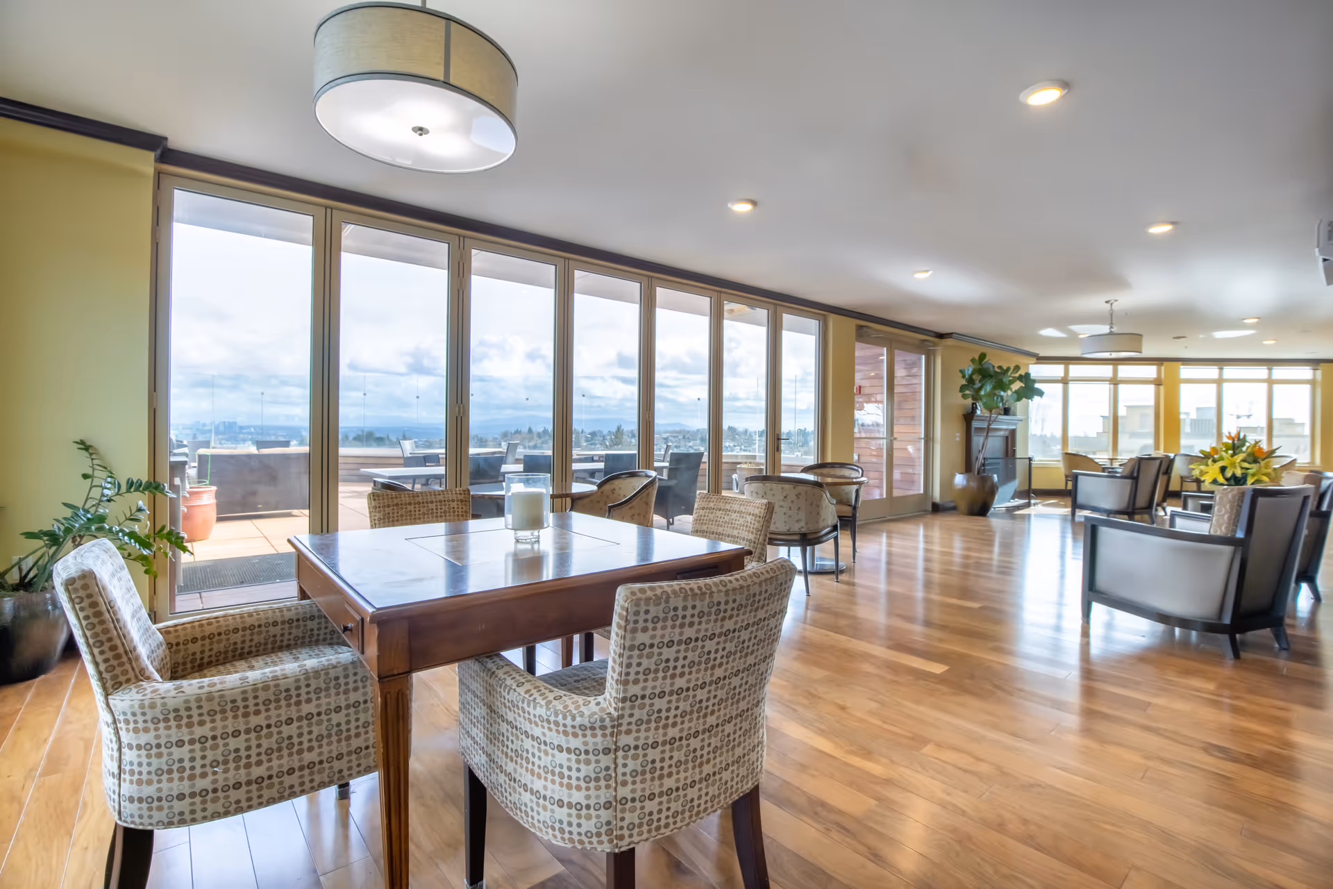 Spacious senior living common room with a wooden dining table, patterned chairs, lounge seating, and large floor-to-ceiling windows overlooking a terrace.