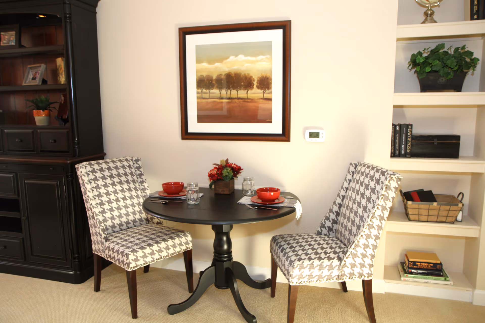 A small round black dining table set for two with red bowls, glasses, and napkins. Two upholstered chairs with a black and white houndstooth pattern are placed on either side of the table. Behind the table is a wall with a framed painting of trees and a thermostat. To the left is a dark wooden cabinet with shelves and drawers, and to the right is a white built-in shelving unit with books, a basket, and a potted plant.