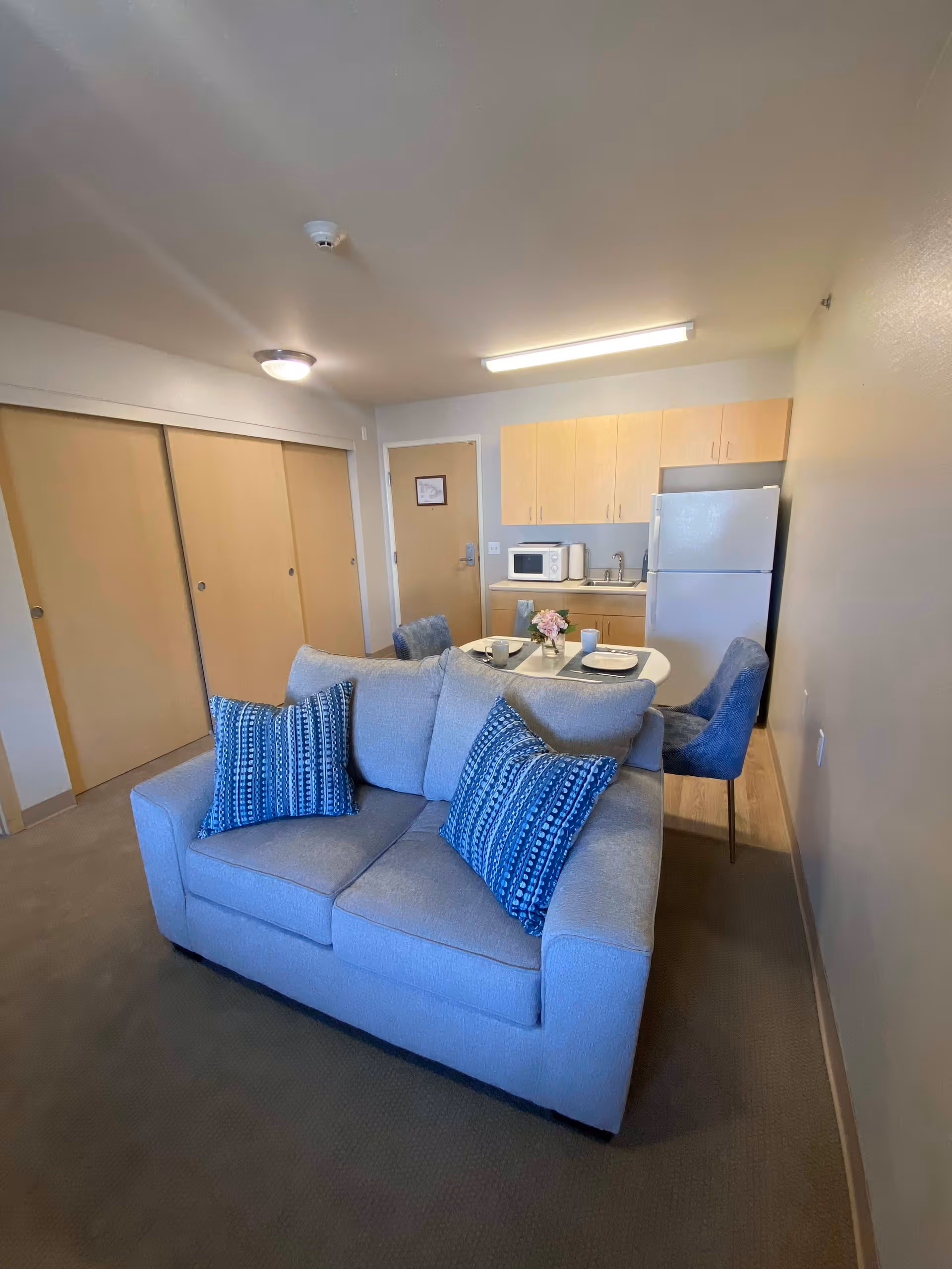 A small living area in a senior living facility featuring a light gray loveseat with two blue patterned pillows. Behind the loveseat is a small dining table set with plates, cups, and a flower centerpiece. The kitchenette includes light wood cabinets, a white refrigerator, a microwave, and a sink. There are sliding closet doors and a beige carpeted floor.