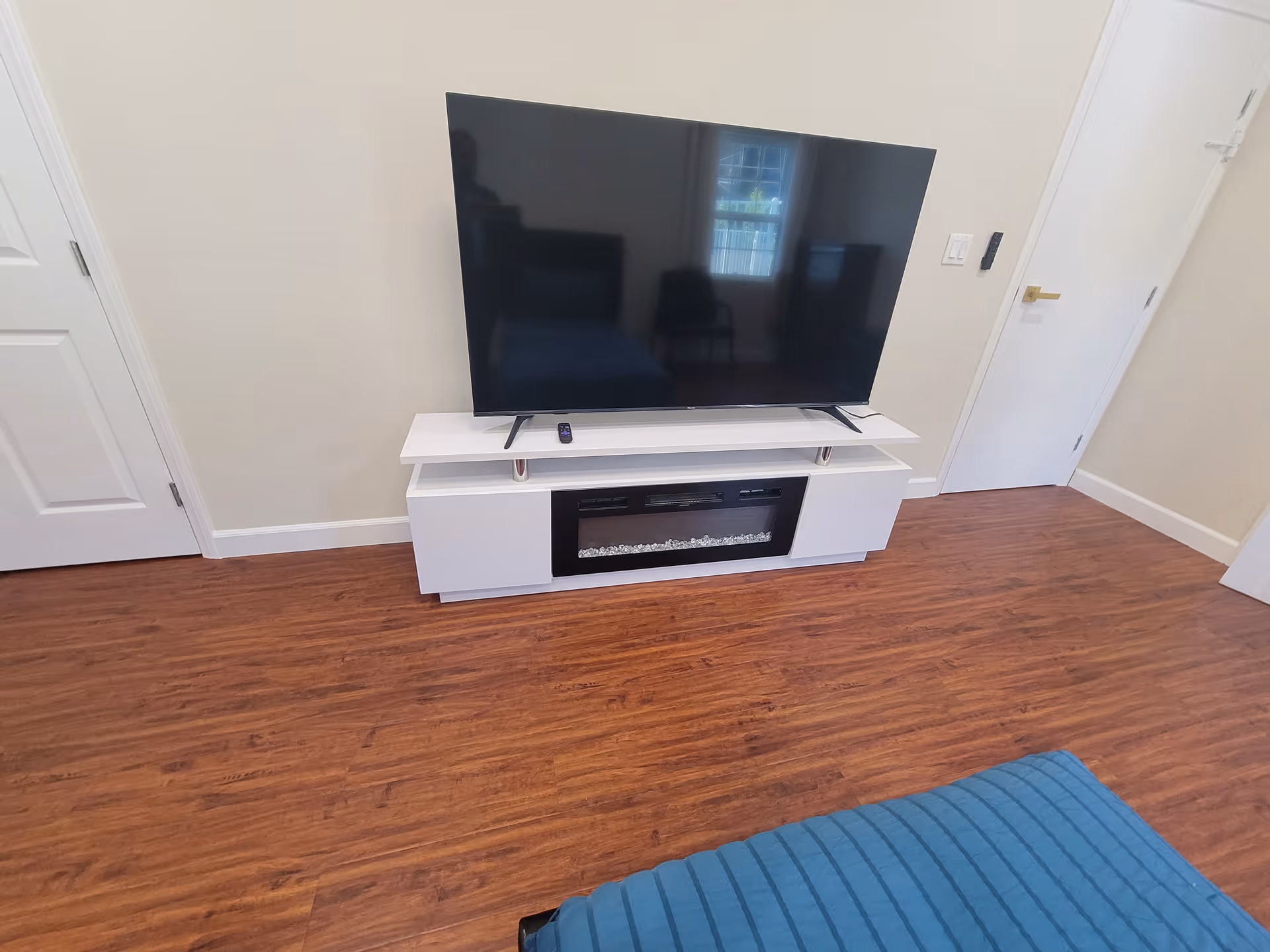 A modern living room area with a large flat-screen TV placed on a white TV stand that includes an electric fireplace. The room has wooden flooring, beige walls, a white door on the right, and a blue cushioned piece of furniture partially visible in the foreground.