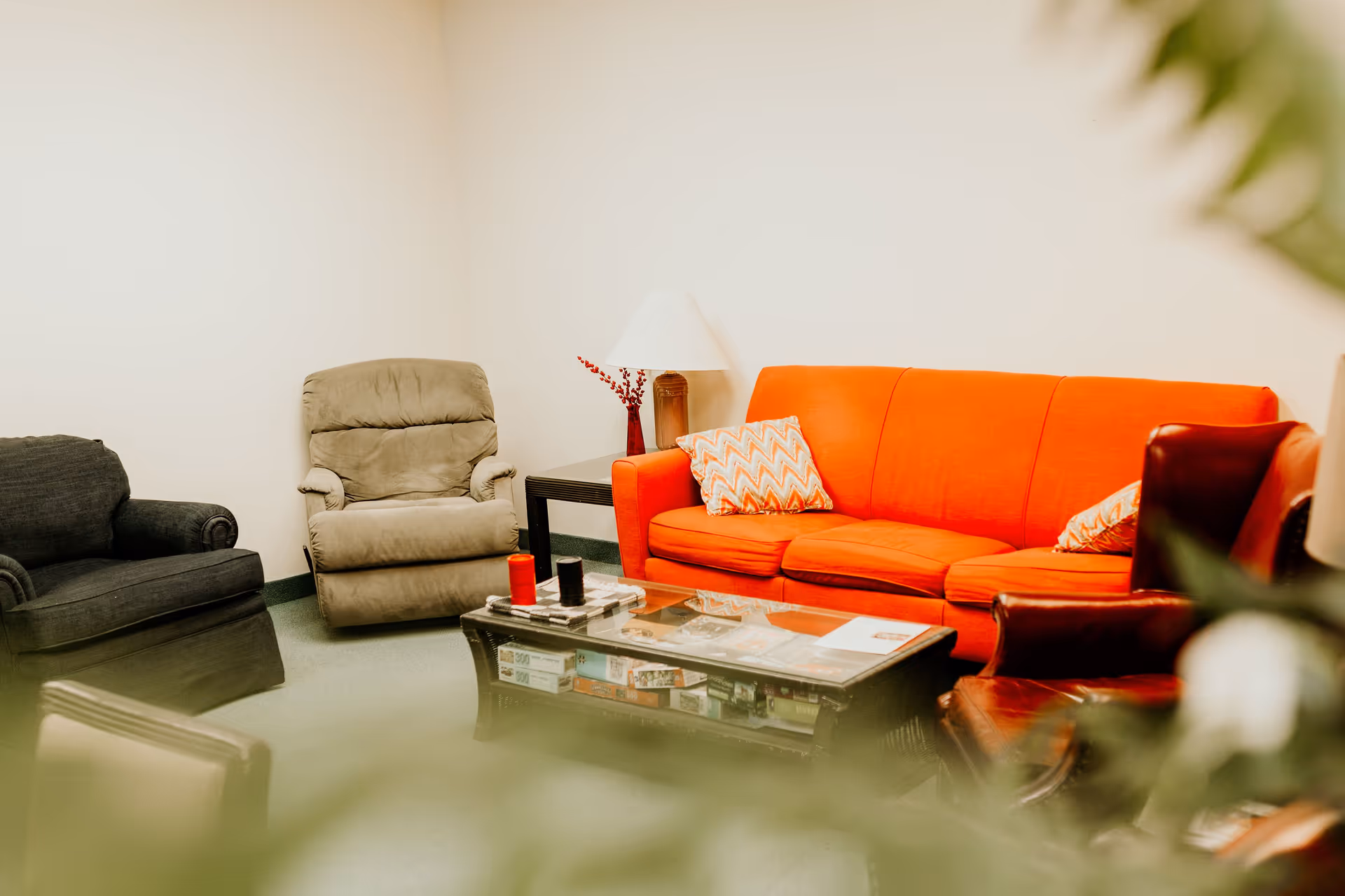 A cozy living room with a bright orange sofa adorned with patterned cushions, a beige recliner chair, a dark gray armchair, and a dark wooden coffee table with board games underneath. A side table with a lamp and a red vase with decorative branches is placed next to the sofa. The room has light-colored walls and green carpet flooring, with some blurred greenery in the foreground.