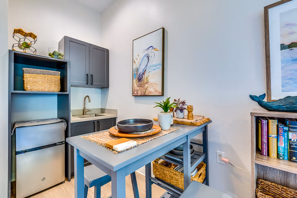 Small kitchenette with a blue island table set with bowls and plants, gray cabinets, a mini fridge and sink, and coastal artwork on the wall.