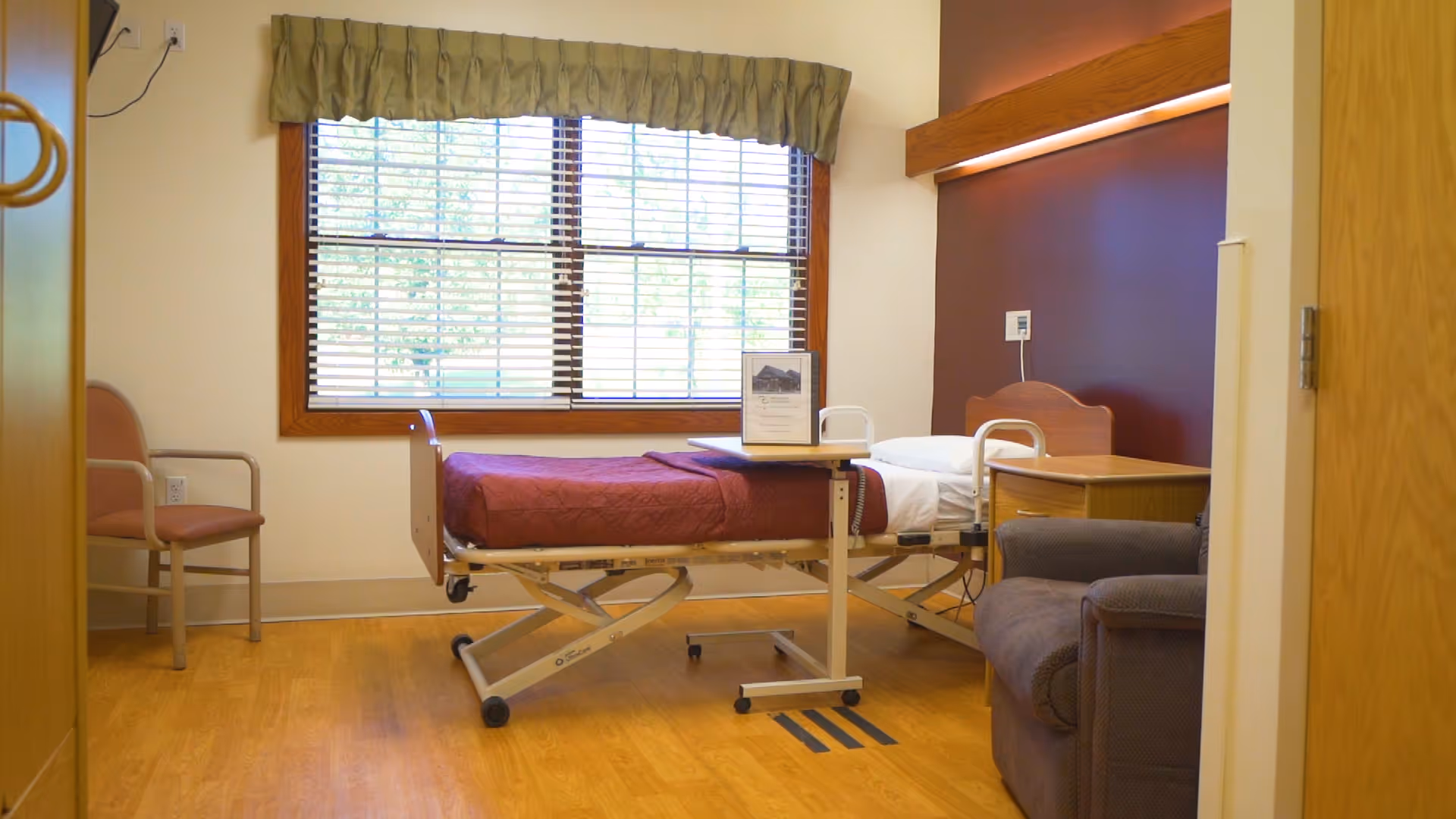 A senior living facility bedroom with a hospital-style bed covered with a maroon blanket, a wooden nightstand, a gray armchair, and a chair near the window with blinds and a green valance.
