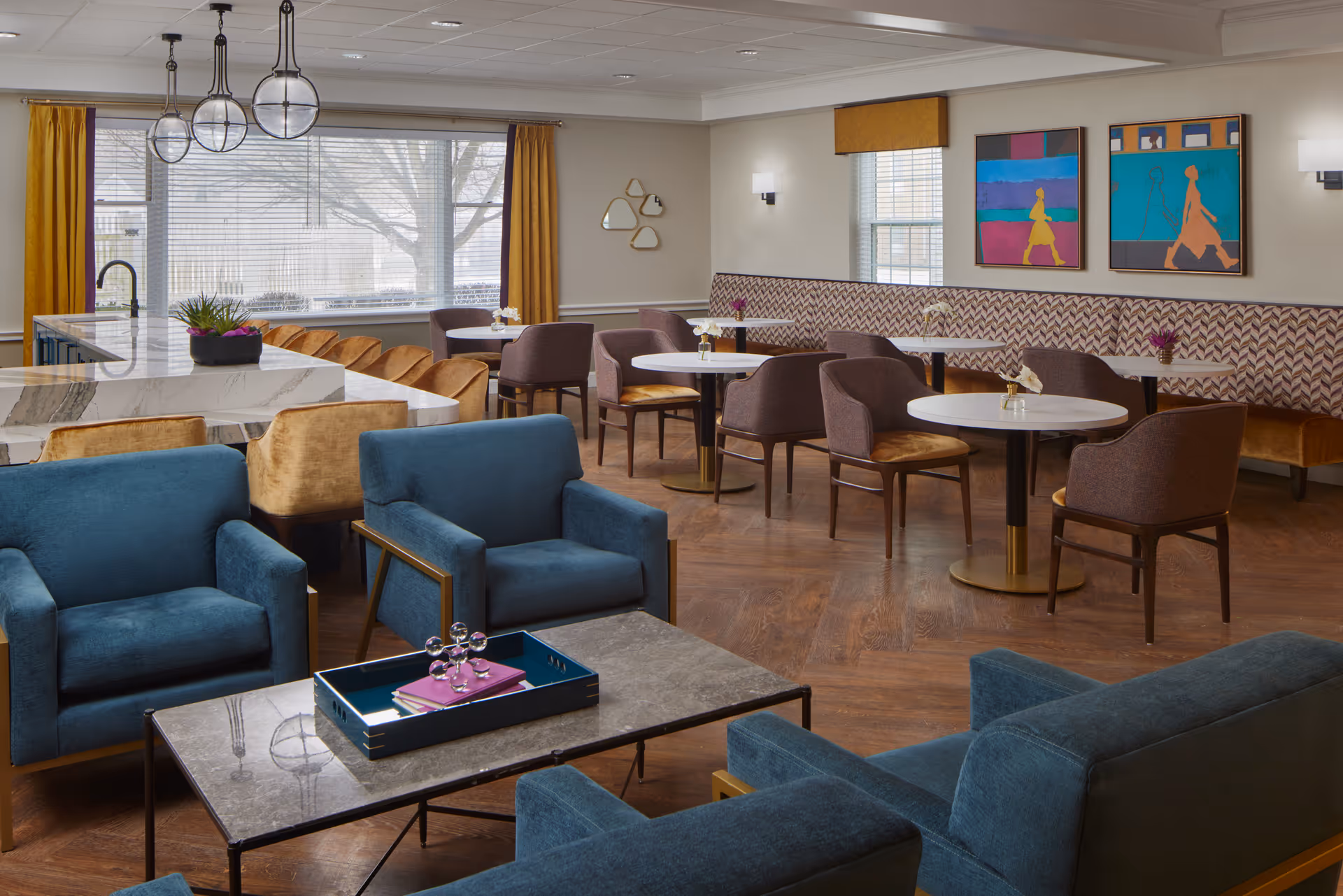 A modern and cozy common area with blue armchairs around a coffee table in the foreground, and several round dining tables with chairs and a cushioned bench along the wall in the background. The room features large windows with yellow curtains, contemporary pendant lights, colorful artwork on the walls, and a marble countertop with bar stools.