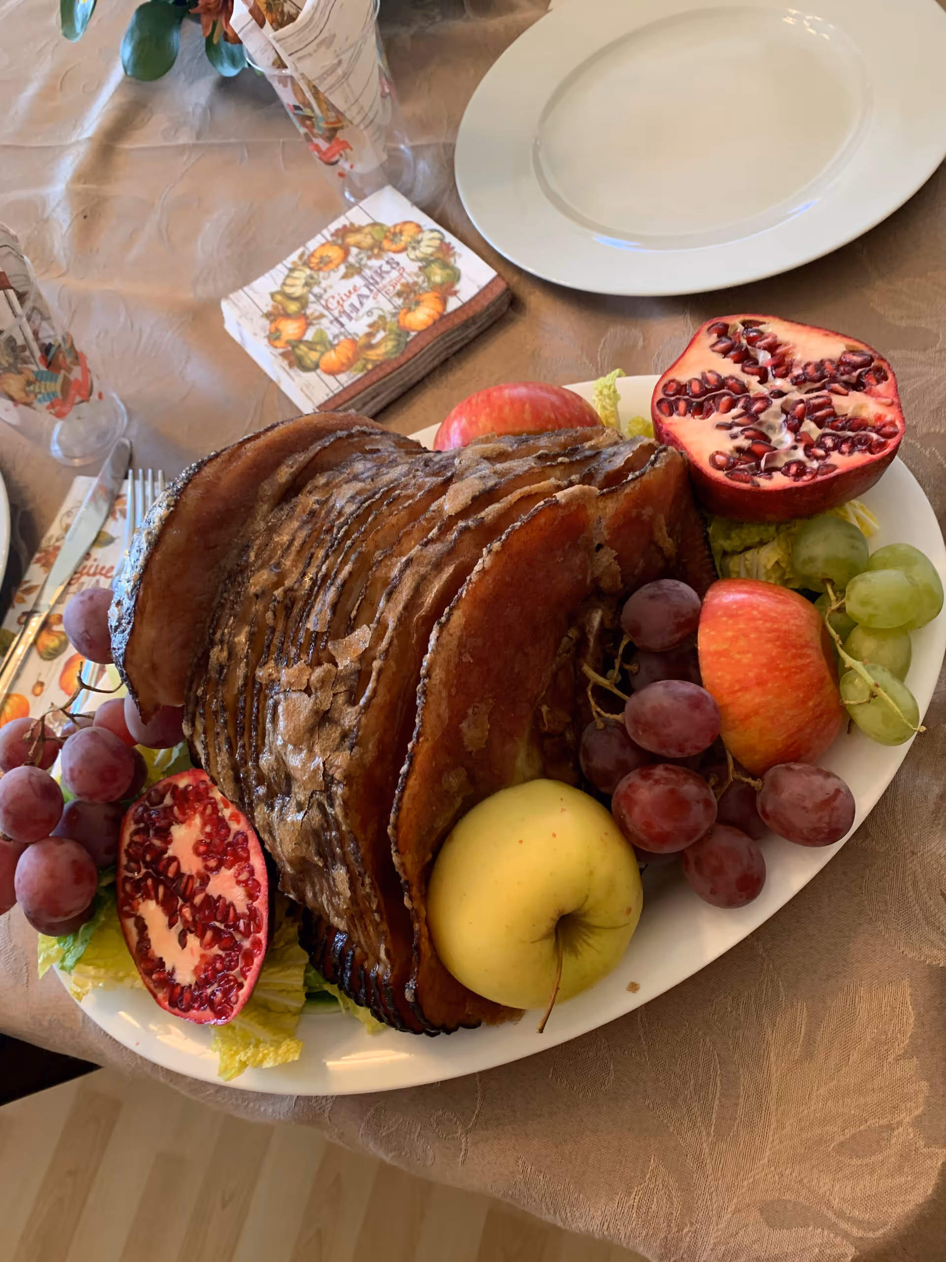 A plate with a large sliced ham surrounded by apples, grapes, and halved pomegranates on a table set with a white plate, utensils, and autumn-themed napkins and cups.