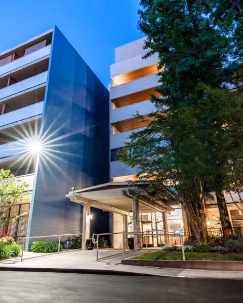 Exterior view of a multi-story building at dusk with illuminated windows and a covered entrance. There are trees and landscaping around the entrance area, and a bright light flare is visible on the left side of the building.