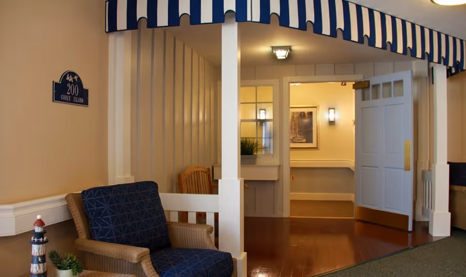 Interior view of a senior living facility hallway with a wicker chair featuring a blue cushion, a small lighthouse decoration, and a potted plant. The hallway has white walls with vertical paneling, a blue and white striped awning above the entrance, and an open door leading to another room with framed artwork and wall sconces.
