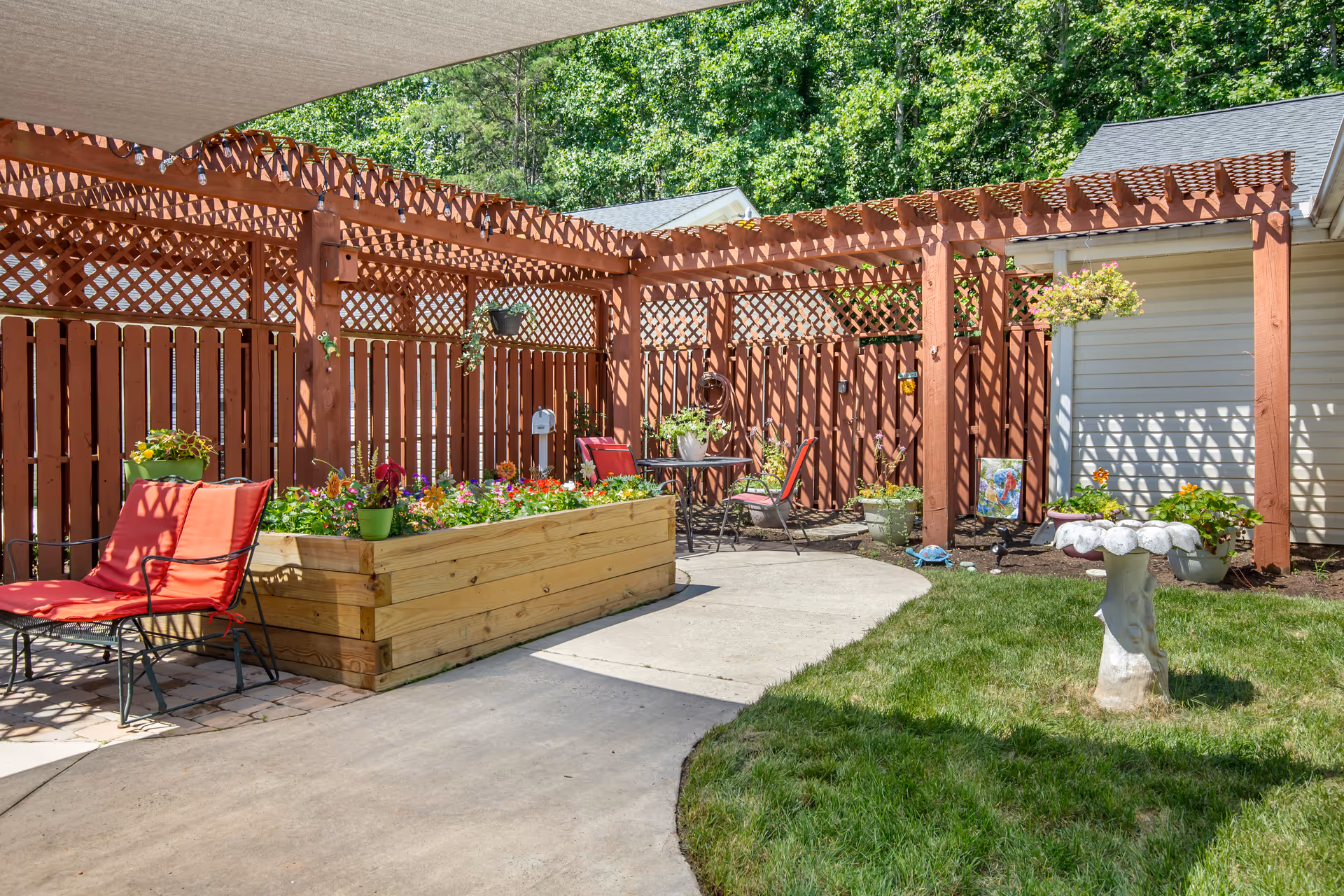 Outdoor patio area with a wooden pergola and lattice fencing. There are red cushioned chairs, a raised garden bed filled with colorful flowers, a small round table with two chairs, hanging flower pots, and a birdbath on a grassy lawn. Trees and a building with beige siding are visible in the background.