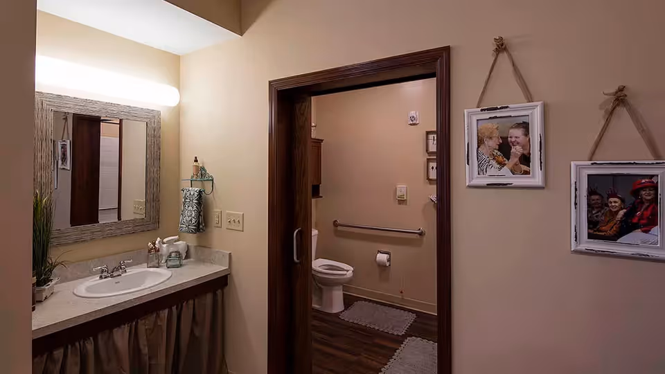 Interior view of a bathroom area in an assisted living facility. The image shows a sink with a large mirror above it on the left side, a towel hanging on a small shelf, and a doorway leading to a toilet with grab bars and a toilet paper holder. The walls are beige, and there are framed photos of elderly people hanging on the wall to the right of the doorway.
