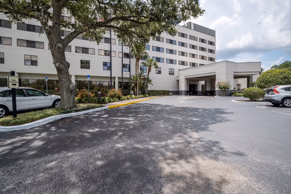 Exterior view of a multi-story senior living facility building with a covered main entrance, surrounded by trees, bushes, and parked cars under a partly cloudy sky.