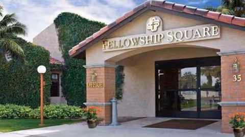 Entrance to Fellowship Square Historic Mesa facility with a covered walkway, glass doors, and a sign above reading 'Fellowship Square'. The building exterior features brick and stucco with greenery and palm trees nearby.