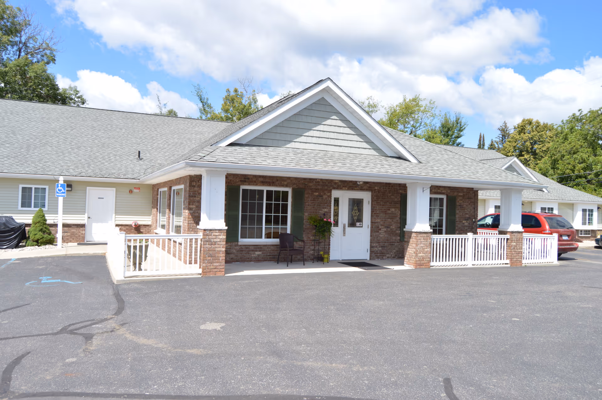Exterior view of a single-story senior living facility building with a covered entrance, brick and siding facade, a white door, windows with green shutters, a chair and potted plant on the porch, and a parking lot with a handicapped parking space in front.
