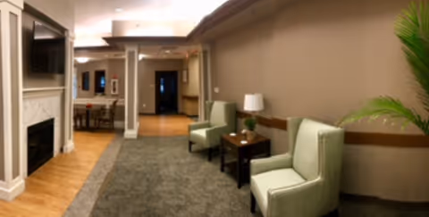 Interior common lounge with two upholstered chairs and a side table in a corridor leading to a dining area, a fireplace with a wall-mounted TV, and a potted plant.