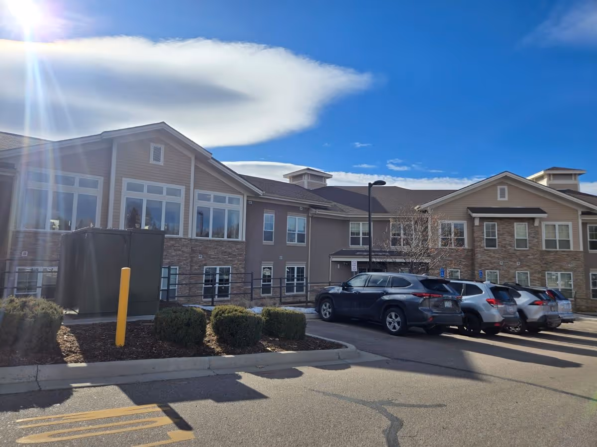 Front exterior of a two-story assisted living building with parked cars in the lot under a bright blue sky.