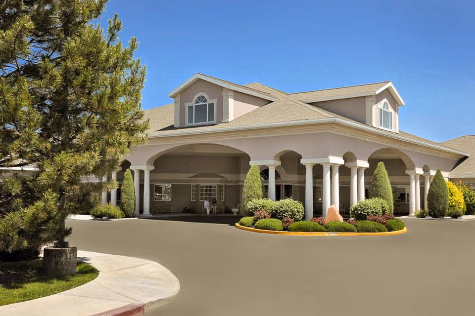 Front exterior view of a senior living facility building with a covered entrance supported by white columns, landscaped bushes and trees, and a clear blue sky.