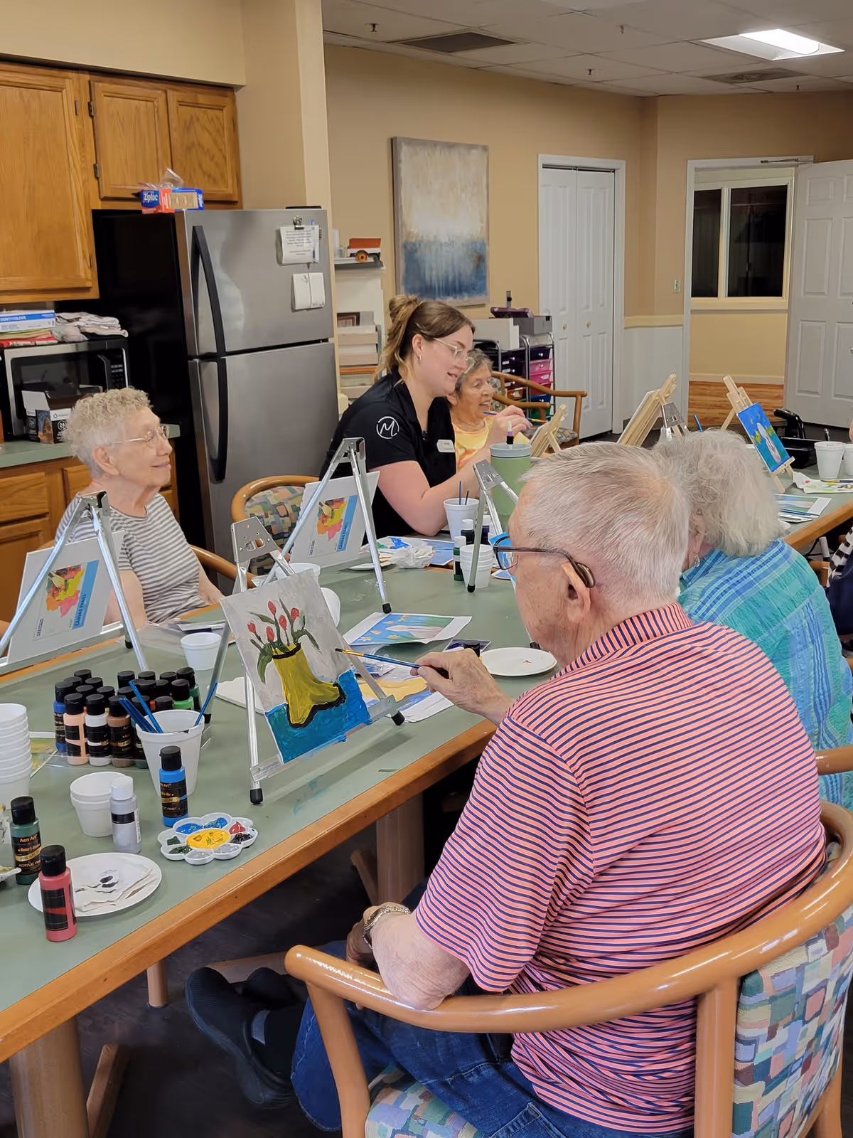 A group of elderly people and a caregiver sitting around a table in a room with wooden cabinets and a refrigerator, engaging in a painting activity with easels, paint bottles, and brushes on the table.