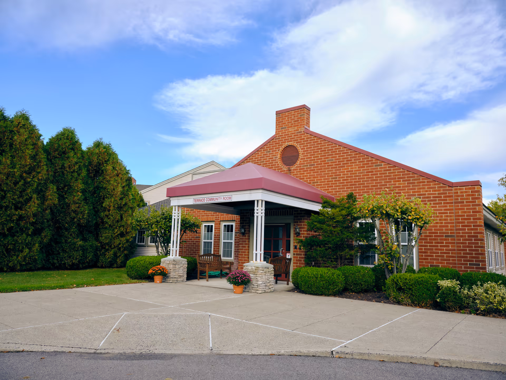 Exterior view of a brick building with a covered entrance labeled 'Terrace Community Room'. The building is surrounded by neatly trimmed bushes, small trees, and potted flowers. The sky is partly cloudy with blue patches.