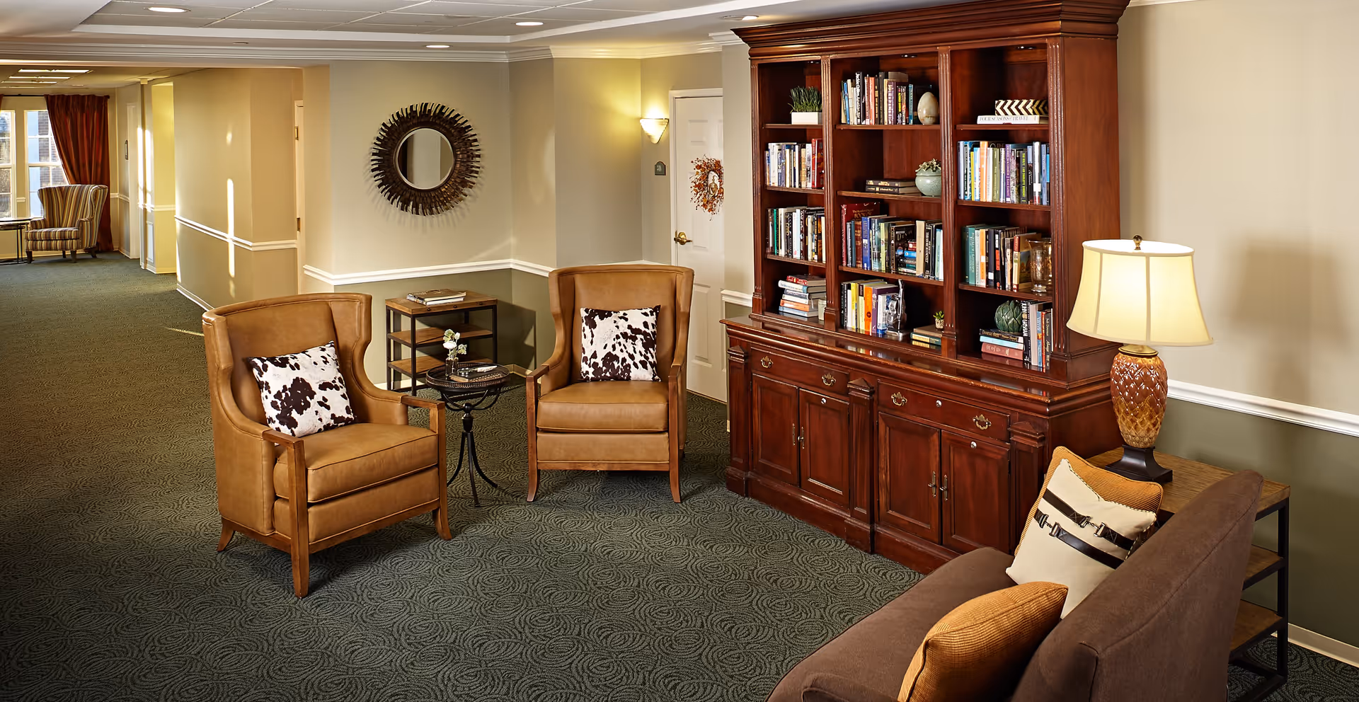A cozy seating area in a senior living facility hallway featuring two brown leather armchairs with patterned pillows, a small round table between them, a large wooden bookshelf filled with books, a brown sofa with cushions, and a table lamp. The hallway has carpeted floors, beige walls with white trim, and a decorative round mirror on the wall.