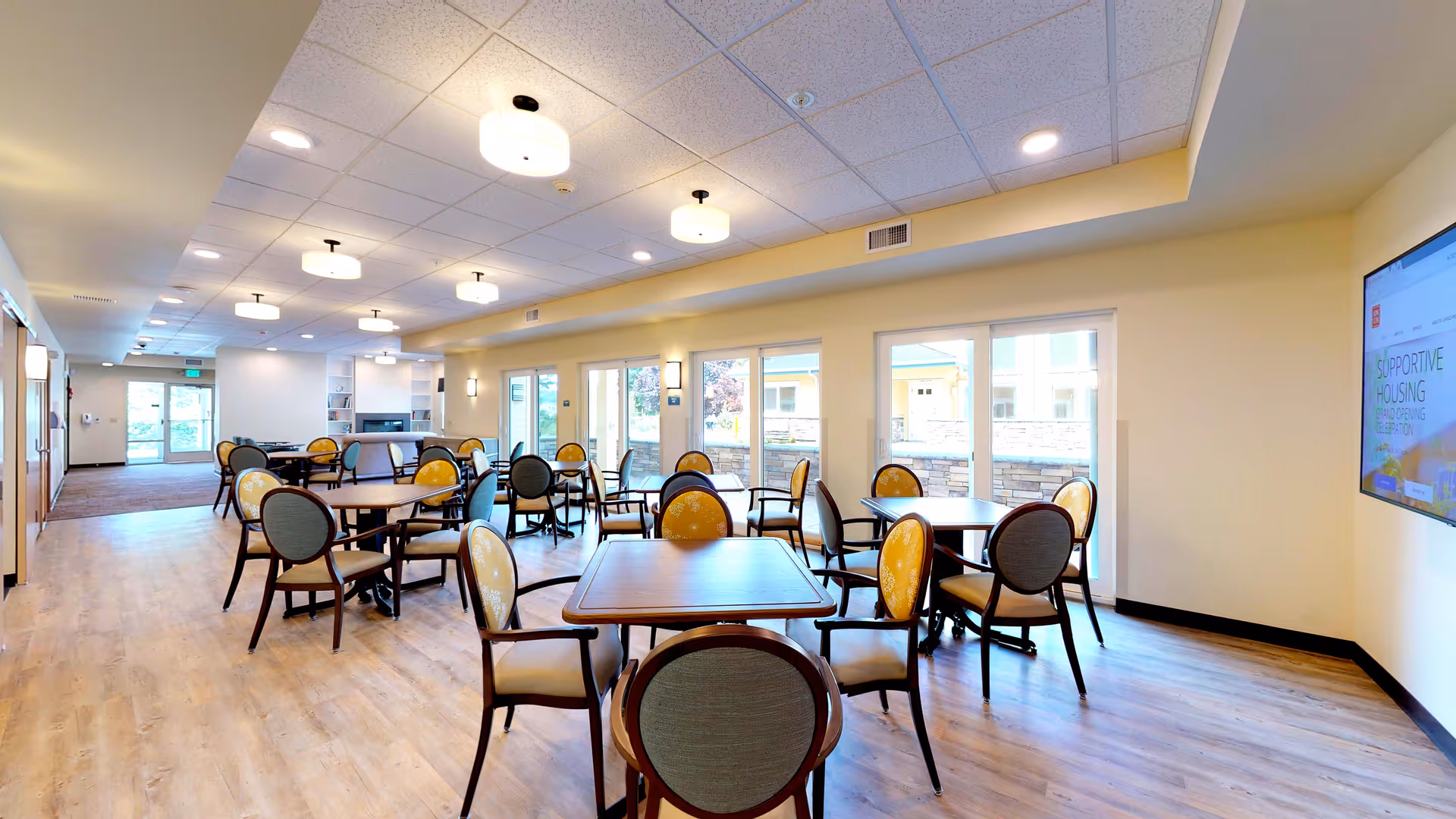 Bright communal dining area with multiple round tables and chairs, large windows, and ceiling lights.