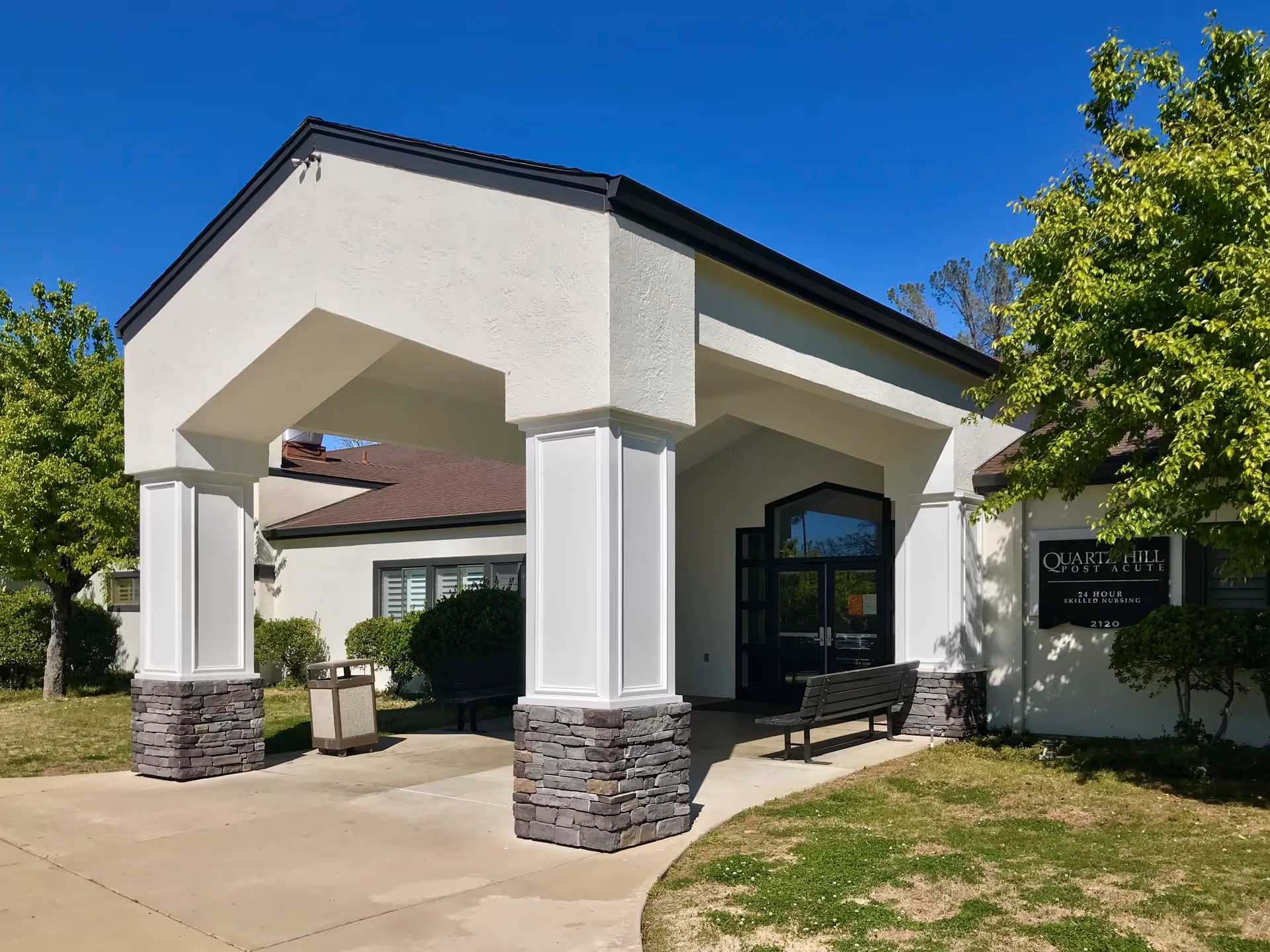 Exterior entrance of Quartz Hill Post Acute facility with a covered walkway supported by white columns with stone bases. There are benches and a trash can near the entrance, surrounded by green trees and bushes under a clear blue sky.