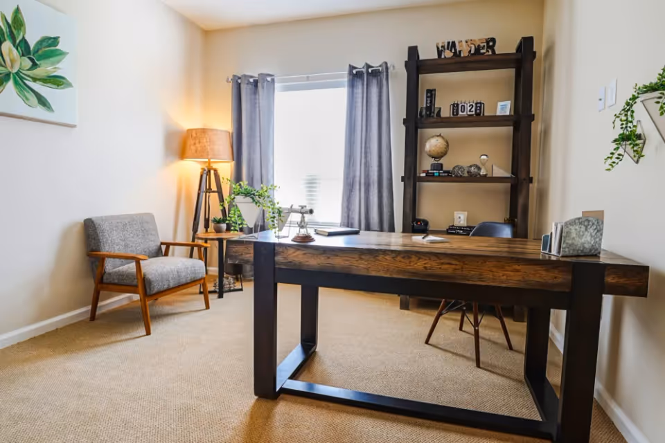 A cozy office space with a large wooden desk, a gray upholstered chair with wooden arms, a floor lamp with a tripod base, a window with gray curtains, and a tall dark wooden bookshelf holding decorative items including a globe and books.