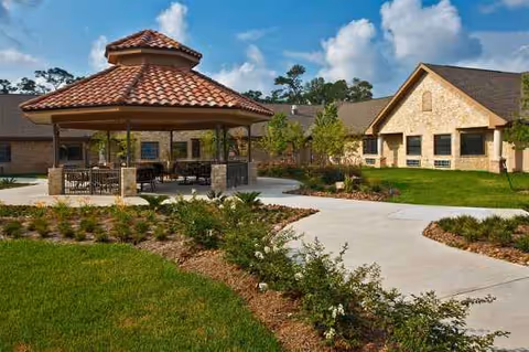 Outdoor courtyard with a shaded pavilion, patio seating, and manicured landscaping beside a stone building