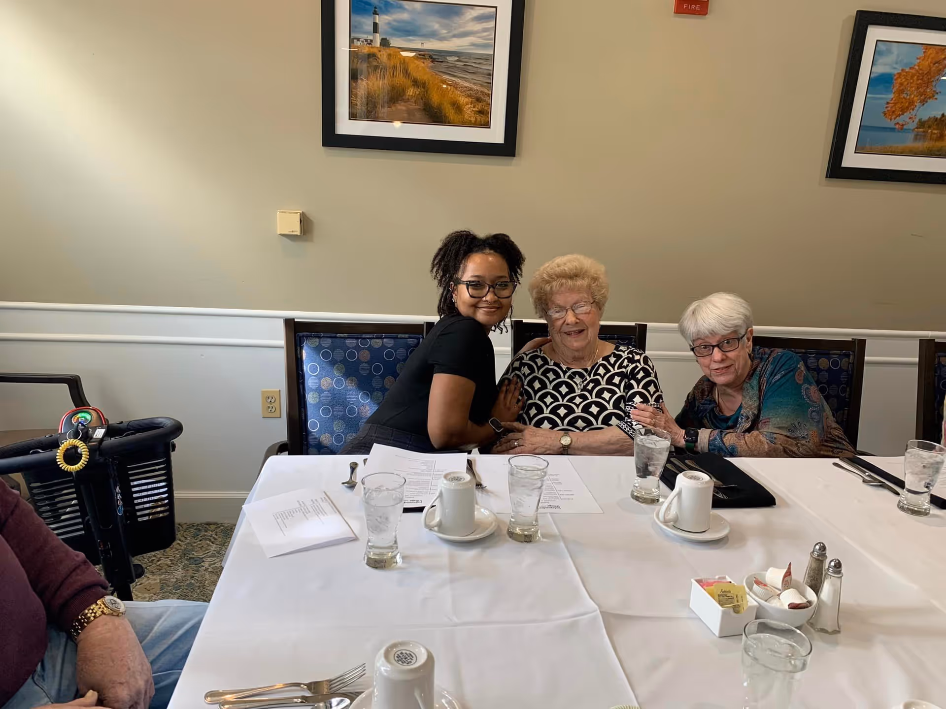 Three women sitting together at a dining table in a room with framed landscape photographs on the wall. The table is set with glasses of water, coffee cups, menus, and condiments. One woman is younger and smiling, while the two older women are seated closely beside her.