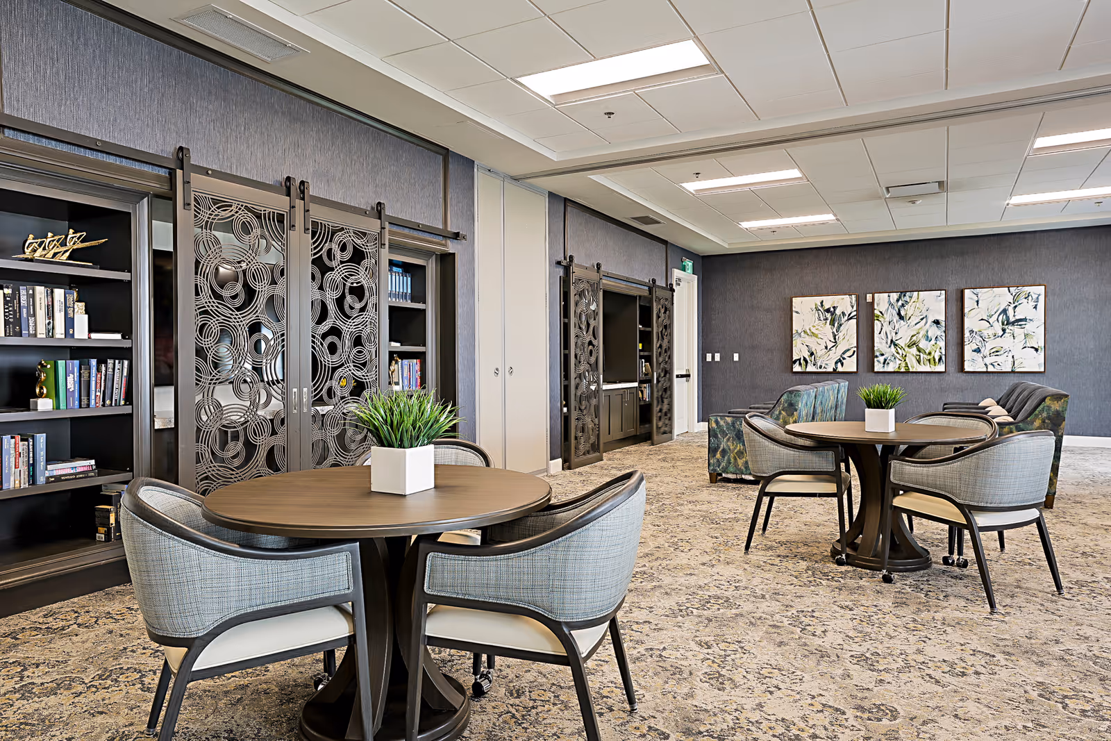 Bright communal lounge with round tables, upholstered chairs, bookshelves and decorative wall art.