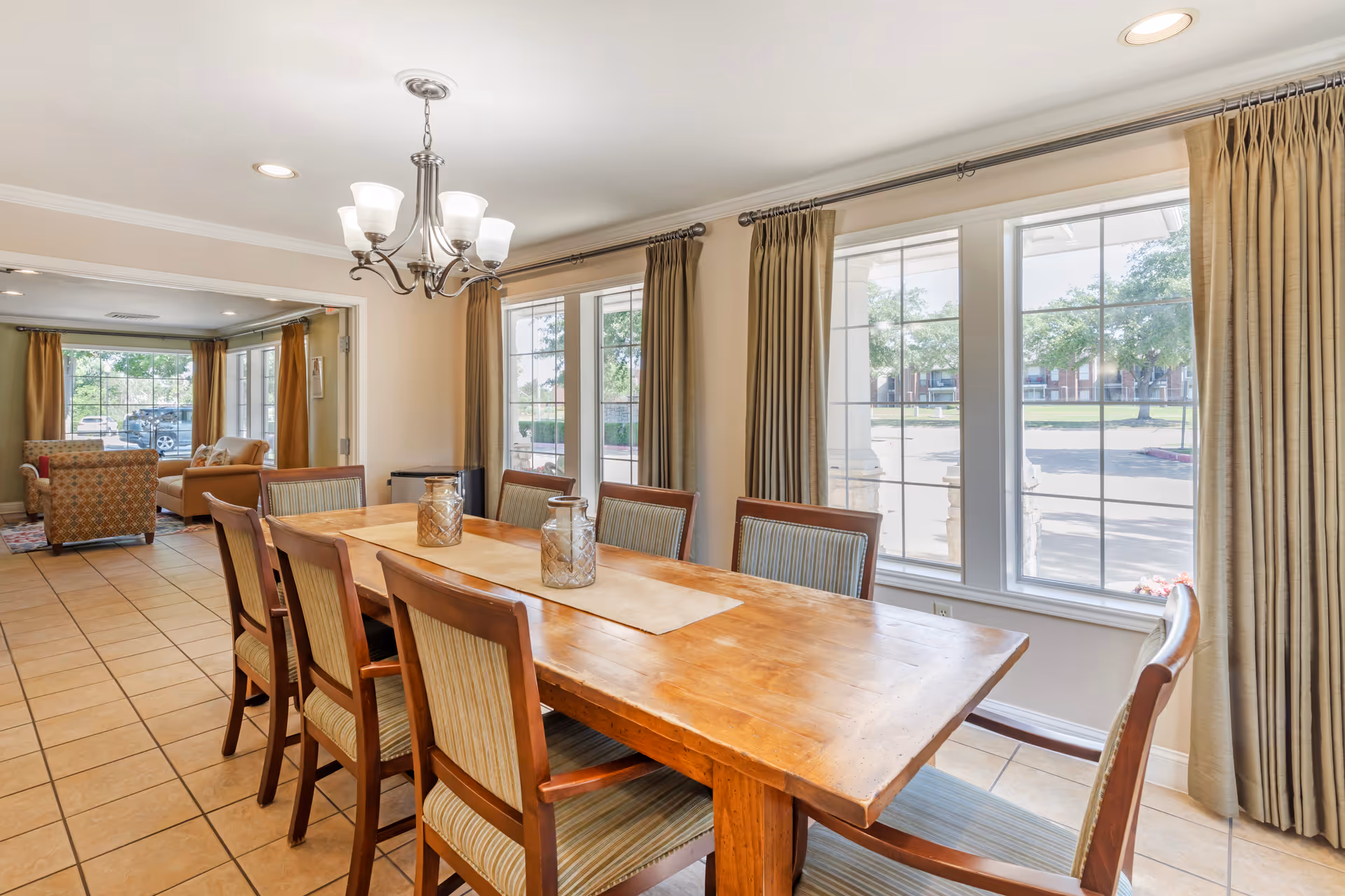 A dining room with a long wooden table surrounded by eight chairs with striped upholstery. Two decorative glass jars are placed on a beige table runner. Large windows with beige curtains allow natural light to fill the room. In the background, there is a sitting area with armchairs and large windows showing an outdoor view.
