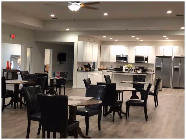 A spacious dining area with several round tables and black cushioned chairs arranged neatly. In the background, there is a modern kitchen with white cabinets, stainless steel appliances including two microwaves and a double-door refrigerator. The room has light-colored wooden flooring and recessed ceiling lights.