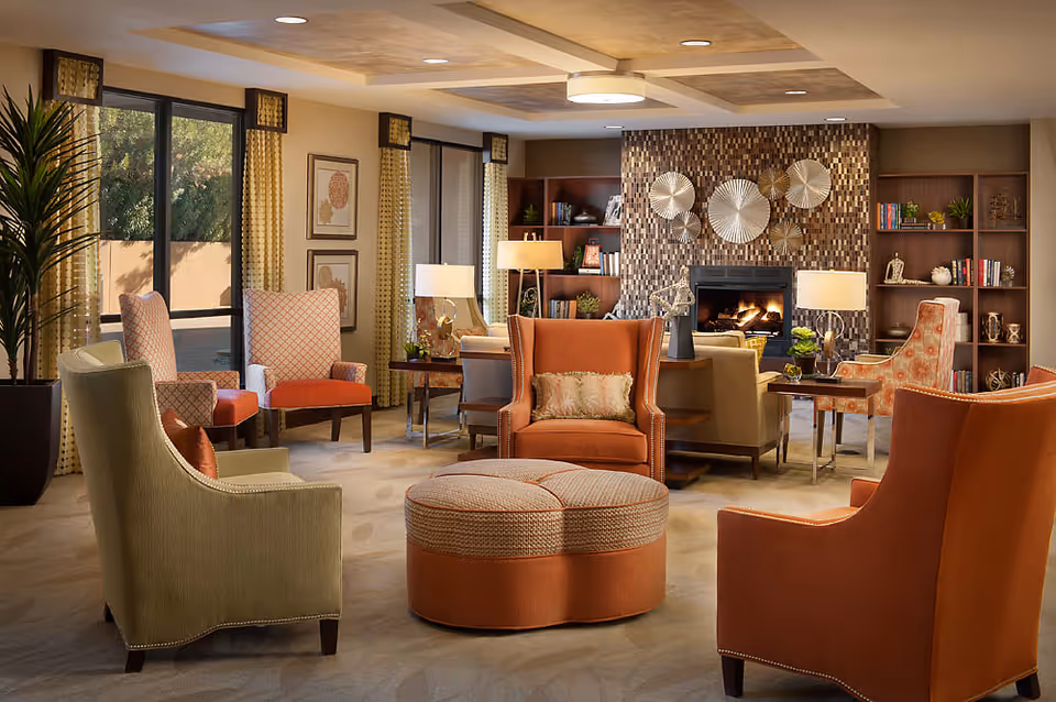 A cozy and well-decorated living room with multiple upholstered armchairs in orange and beige tones arranged around a round ottoman. The room features large windows with yellow curtains, a fireplace with decorative circular wall art above it, built-in bookshelves filled with books and decorative items, and several table lamps providing warm lighting.
