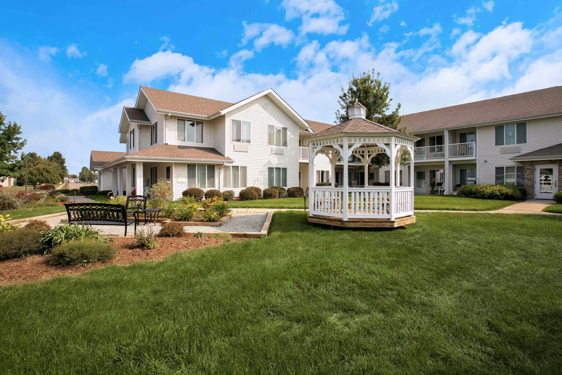 Well-maintained senior living courtyard with a white gazebo, benches, green lawn, and two-story light-colored buildings under a blue sky.