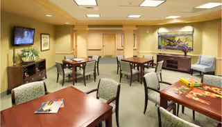 A well-lit common area in a senior living facility with several wooden tables and chairs arranged for group activities. One table has a puzzle in progress, and another has magazines. A flat-screen TV is mounted on the wall, and there is a large aquarium on a cabinet against the far wall. The room has neutral-colored walls and carpeted flooring.