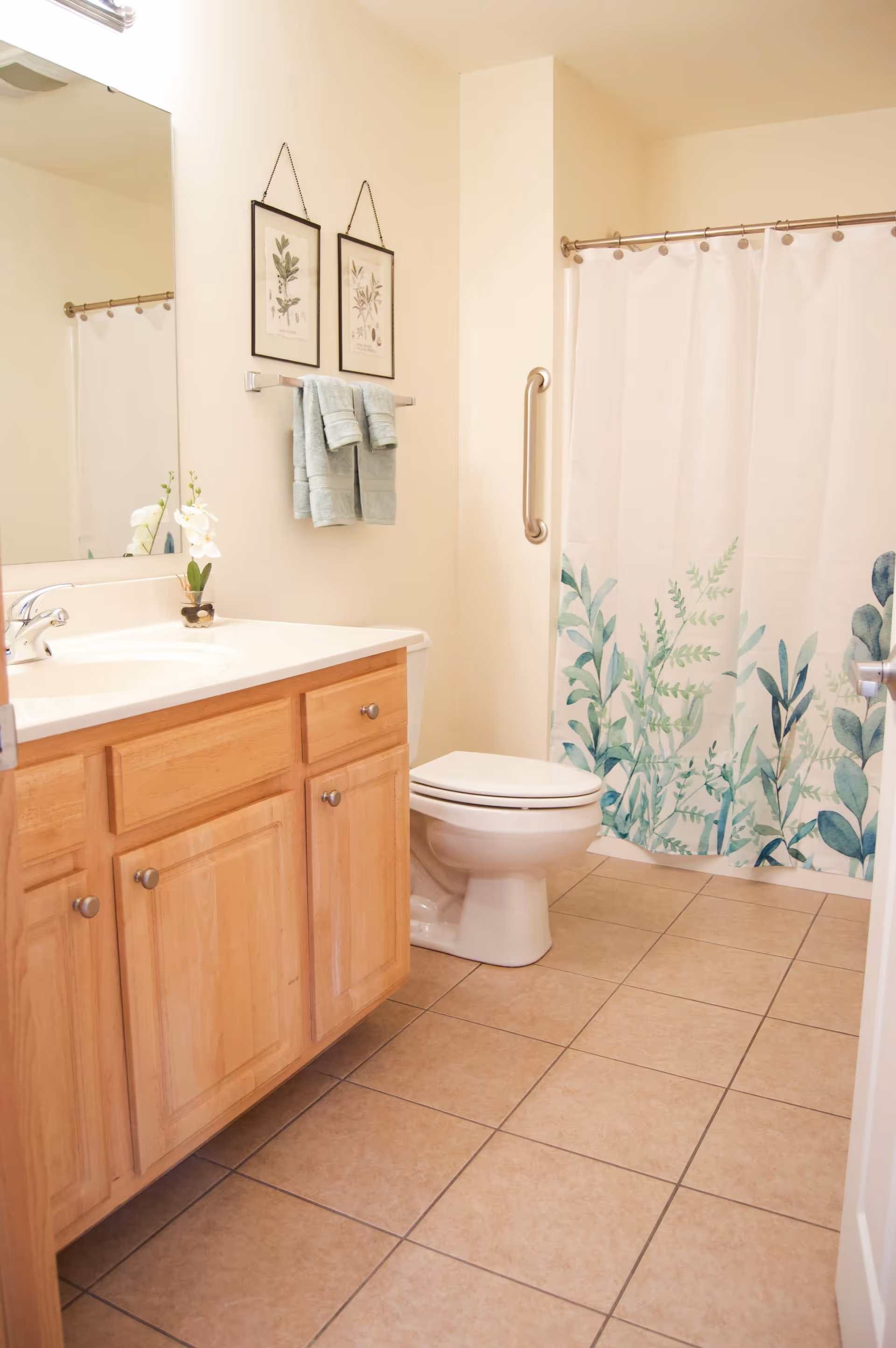 A clean bathroom with a wooden vanity cabinet, a white sink, a toilet, and a shower with a white curtain featuring green leafy patterns. Two framed botanical prints hang on the wall above a towel rack with folded light blue towels. The floor is tiled in a light brown color.