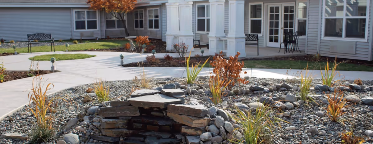 Outdoor courtyard area of a senior living facility with a rock garden featuring stacked stones and plants, surrounded by paved walkways, benches, and a building with multiple windows and a covered porch.