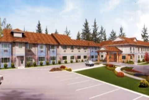 Exterior view of a two-story assisted living facility with a red-tiled roof, multiple windows, and a covered entrance. There is a parking lot in front with marked parking spaces and landscaped areas with grass and shrubs.