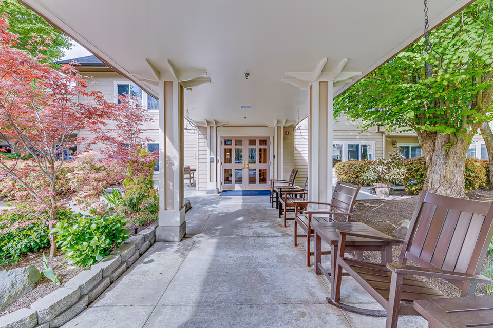 Covered entrance walkway to a senior living building with wooden chairs, columns, and landscaped plantings.