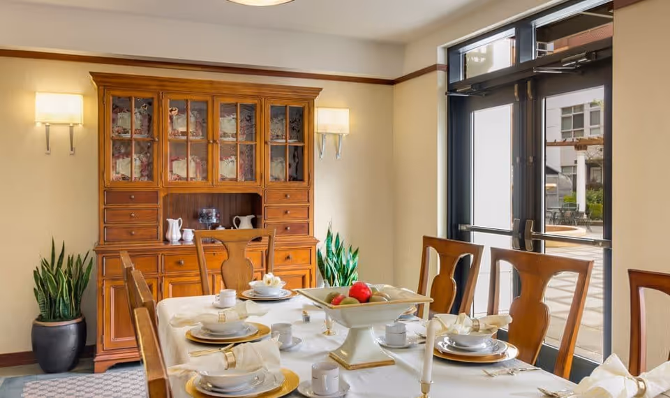 A dining room with a wooden dining table set for a meal, including plates, cups, napkins, and a centerpiece bowl with fruit. The room features wooden chairs, a wooden cabinet with glass doors displaying dishes, two wall-mounted lamps, potted plants, and large glass doors leading outside.