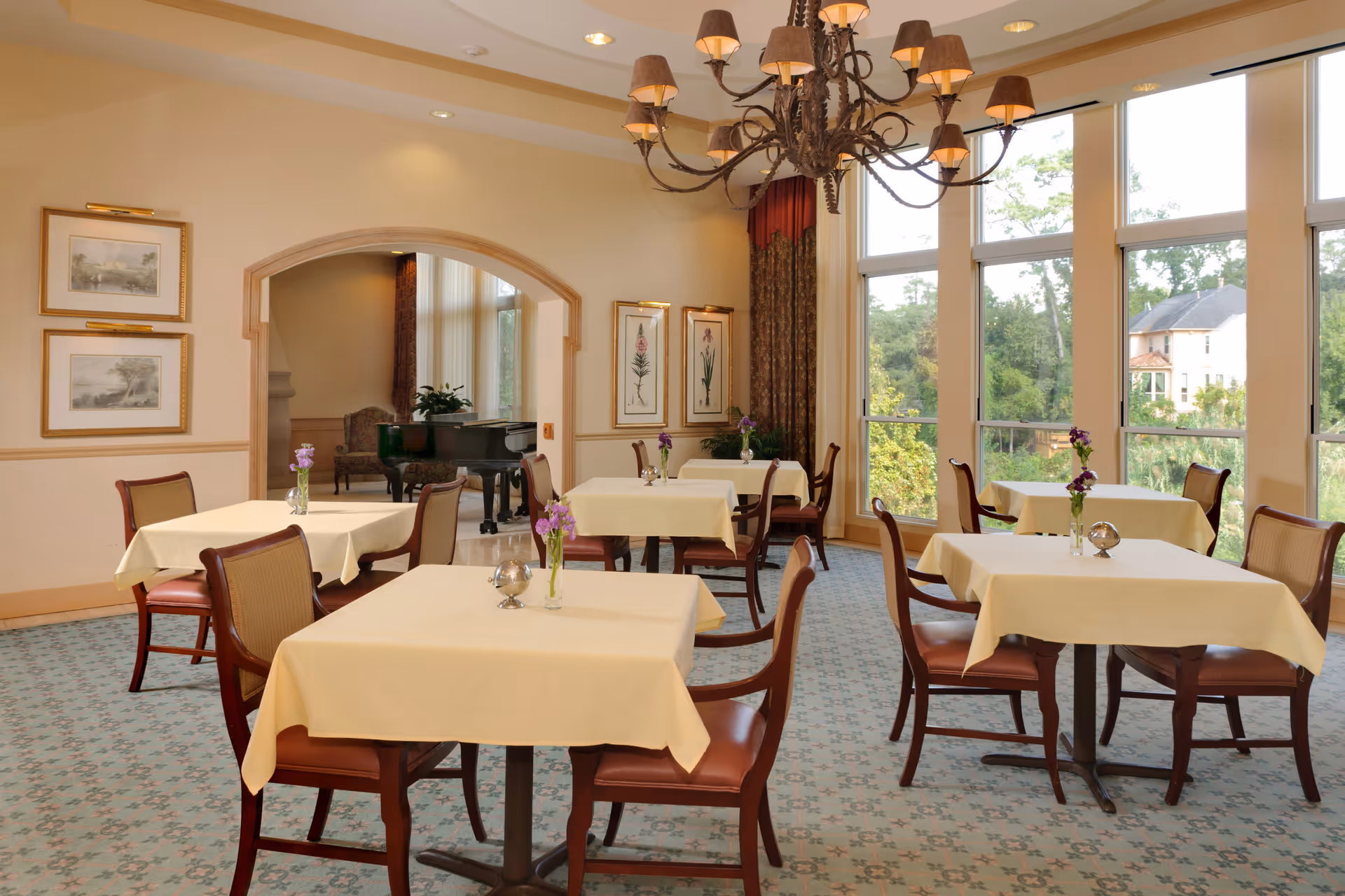 A bright dining room with several tables covered in cream-colored tablecloths, each with a small vase of purple flowers. The room features large windows letting in natural light and offering a view of greenery outside. There is a decorative chandelier hanging from the ceiling and framed artwork on the walls. In the background, a grand piano is visible through an arched doorway.