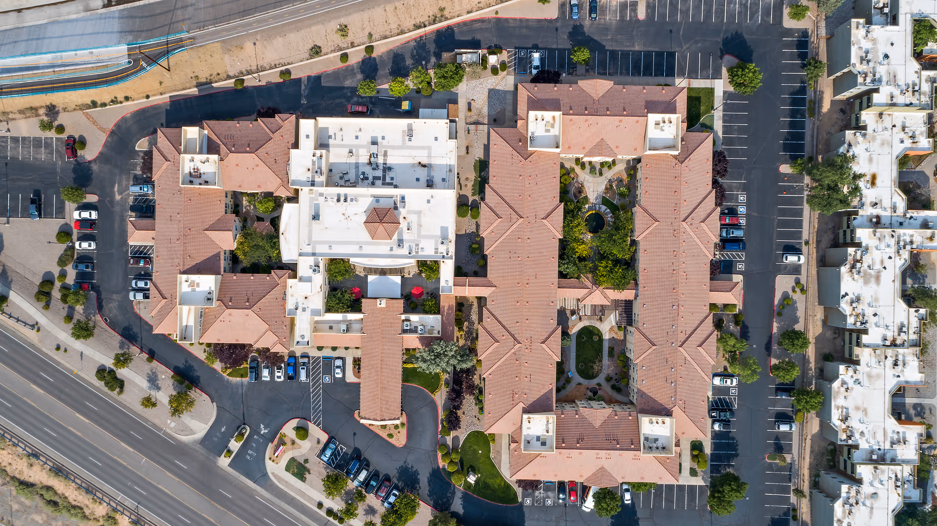 Aerial view of Morada Albuquerque senior living facility showing multiple buildings with red-tiled roofs, parking lots with cars, landscaped courtyards, and surrounding roads.
