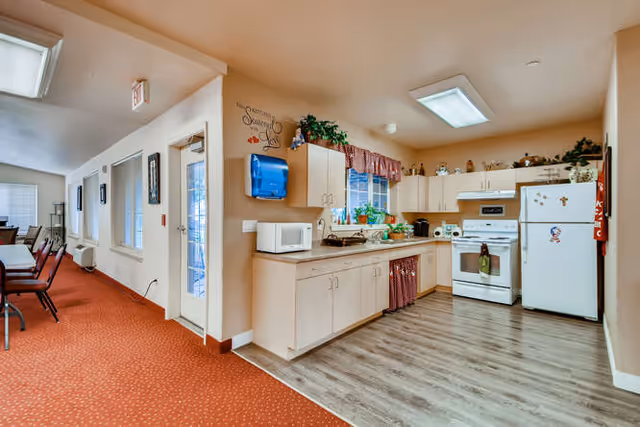 Interior view of a senior living facility showing a kitchen area with white cabinets, a microwave, stove, and refrigerator. The kitchen has a window with a red valance and various plants on the windowsill. Adjacent to the kitchen is a hallway with red carpet and chairs along the wall.