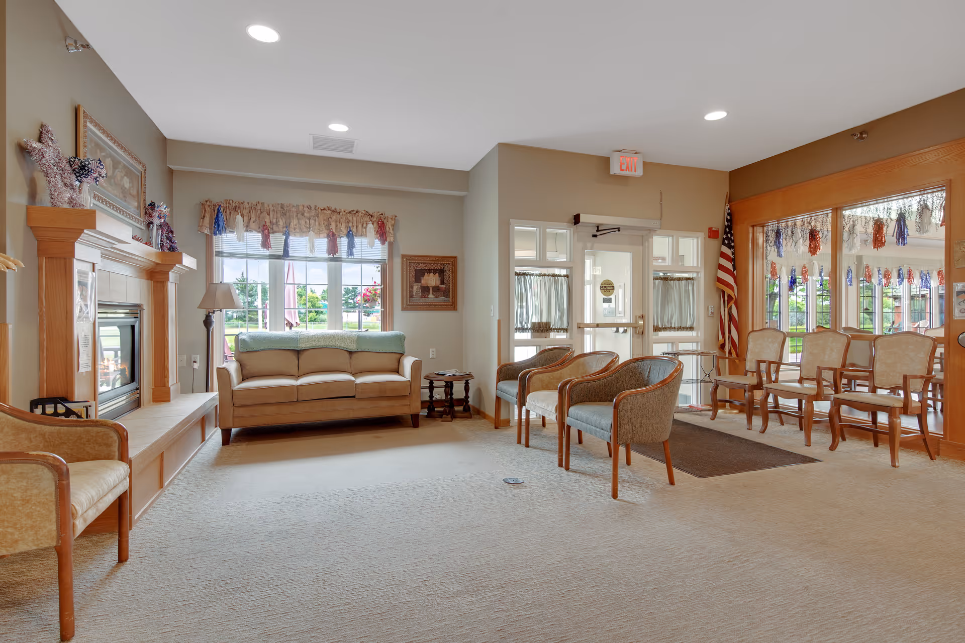 Bright senior-living common lounge with a sofa, armchairs, fireplace, and an entrance with large windows and an American flag.