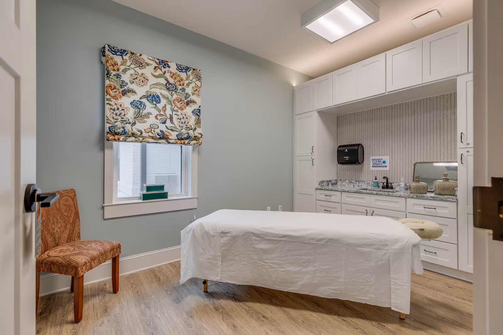 Treatment room with a covered massage table, floral window shade, upholstered chair, and white built-in cabinetry with sink and counter.