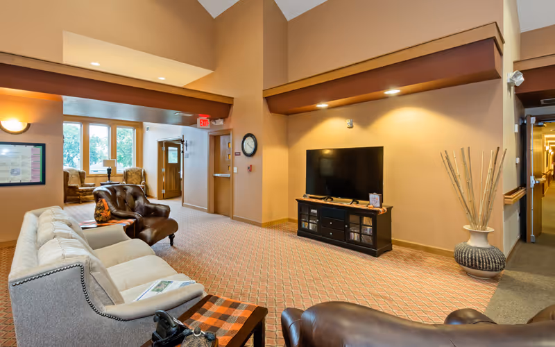 Communal living room area with sofas and armchairs, a TV on a console, and a hallway in an assisted living facility.
