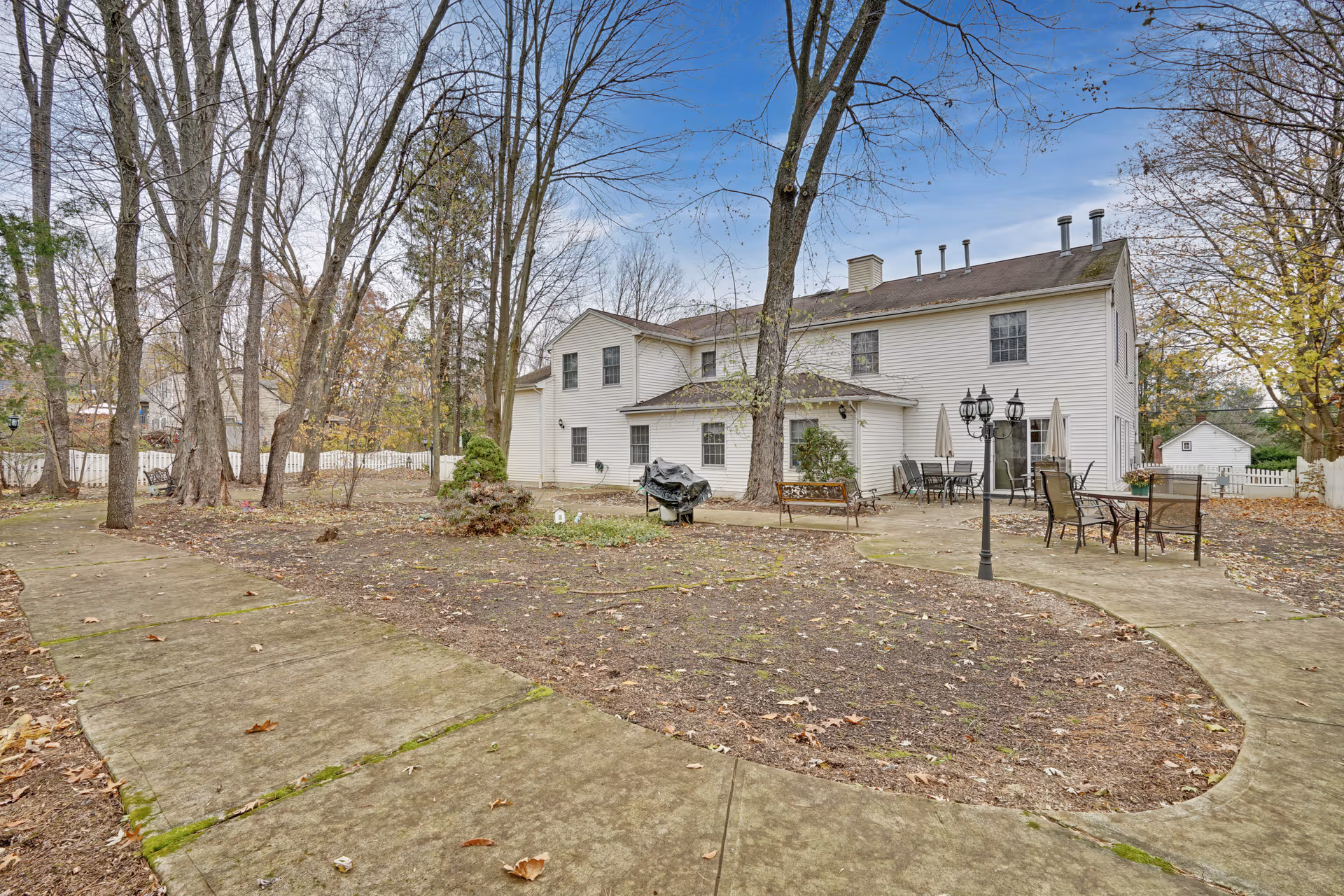 Outdoor view of a two-story white building with a patio area featuring tables, chairs, umbrellas, a bench, and a barbecue grill. The surrounding area has leafless trees and a concrete walkway curving through a yard with sparse grass and fallen leaves under a partly cloudy sky.