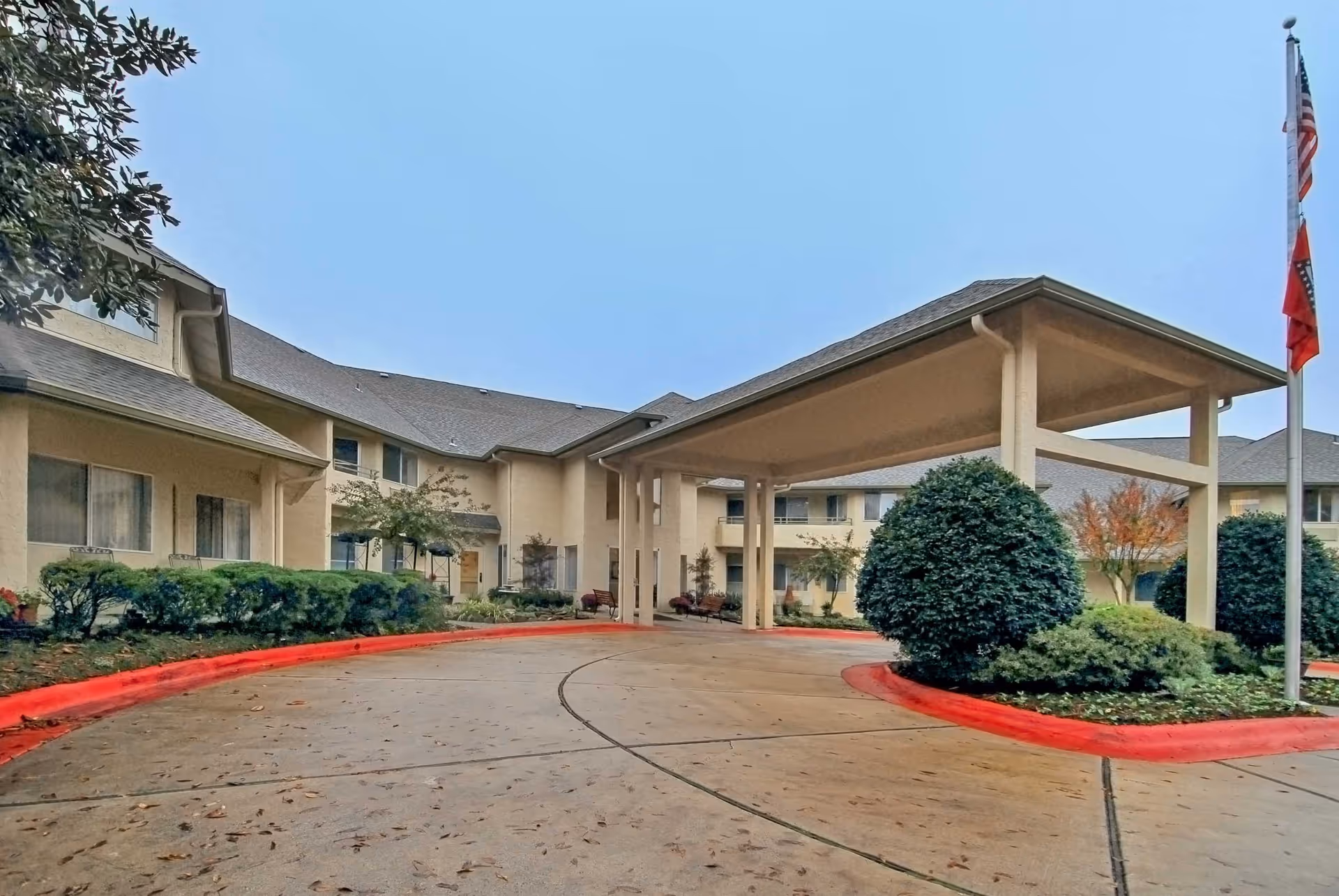 Exterior view of Holiday Andover Place senior living facility showing a covered entrance with a driveway, landscaped bushes, and an American flag on a flagpole under a clear sky.